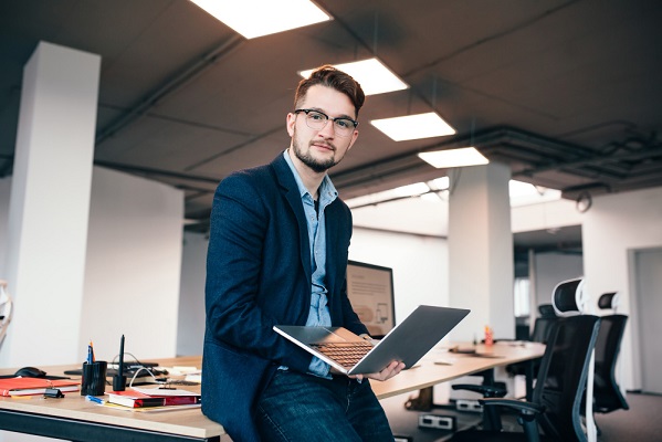 attractive-man-glassess-is-sitting-near-workplace-office-he-wears-blue-shirt-dark-jacket-he-holds-laptop-looks-camera.jpg